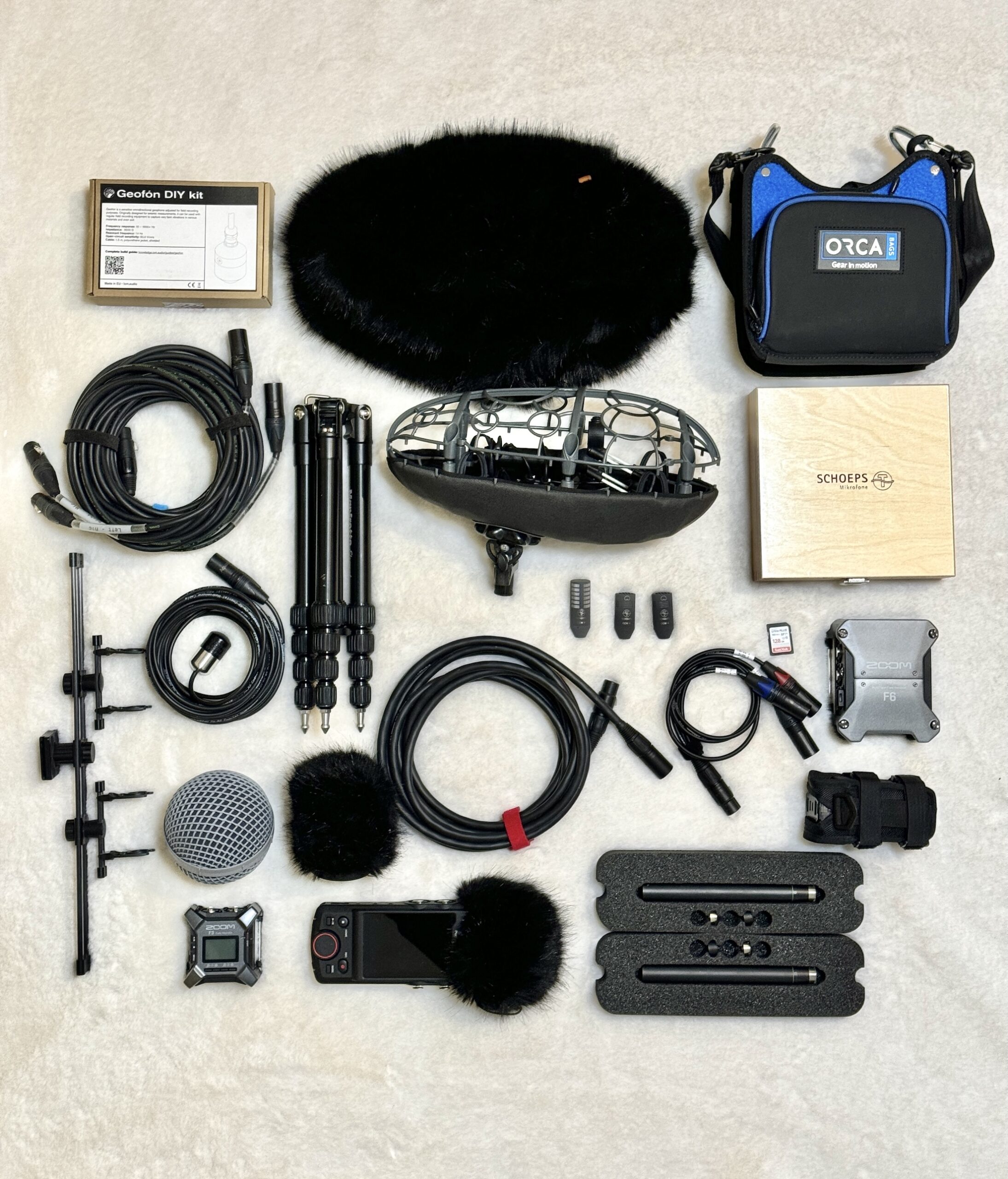 Field recording kit laid out on a pale surface: a blimp windshield and furry cover, shotgun and stereo microphones, portable recorders, a tripod, cables, and carrying cases